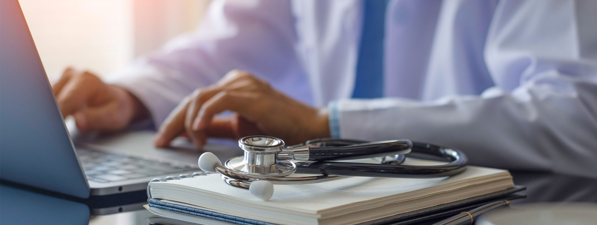 Female doctor in white lab coat typing on laptop computer with notebook and medical stethoscope on the desk at workplace. 