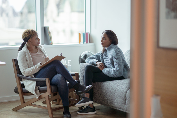 A Therapist meets with her female client in her office. The client is seated on a sofa with her arms across her body as she looks visibly nervous. The Therapist is seated in a chair in front of her as she talks about what to expect from the appointment and takes notes on her clipboard.