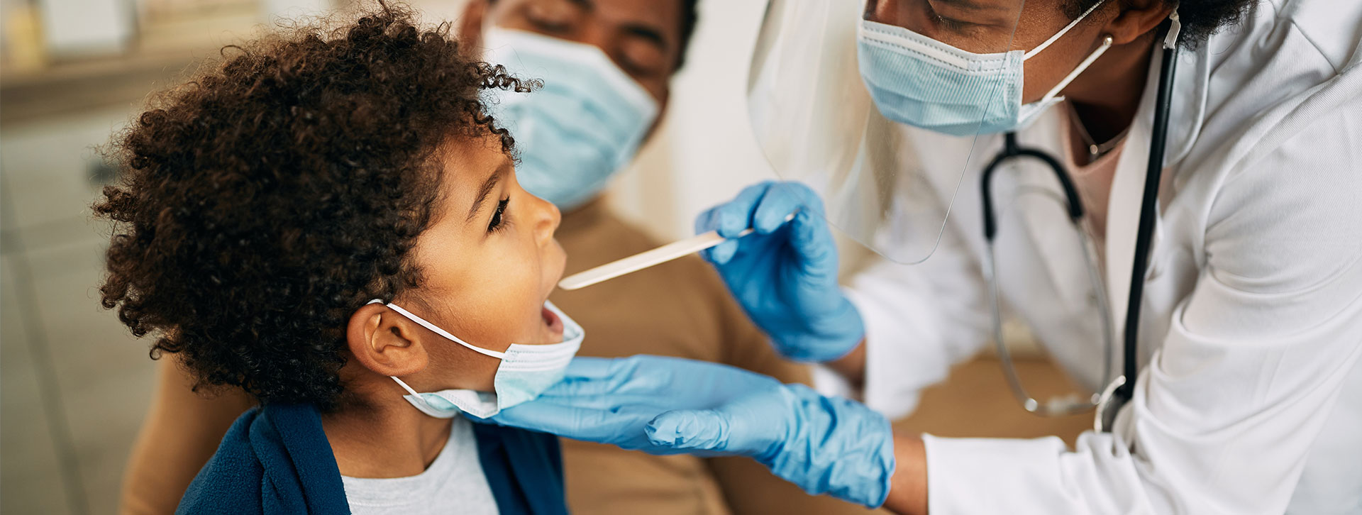 African American doctor with face mask examining boy's throat during a home visit.