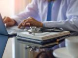 Female doctor in white lab coat typing on laptop computer with notebook and medical stethoscope on the desk at workplace. 