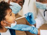 African American doctor with face mask examining boy's throat during a home visit.