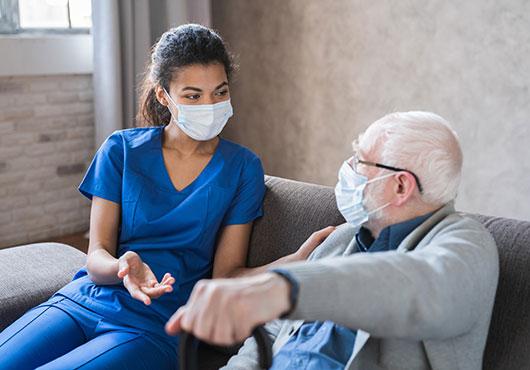 Doctor or female caregiver with senior man grandfather wearing protective mask to protect from Covid 19 at home or nursing home. Doctor talking consulting elderly patient at home