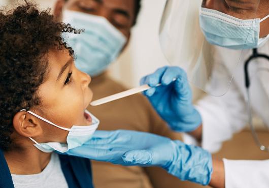 African American doctor with face mask examining boy's throat during a home visit.