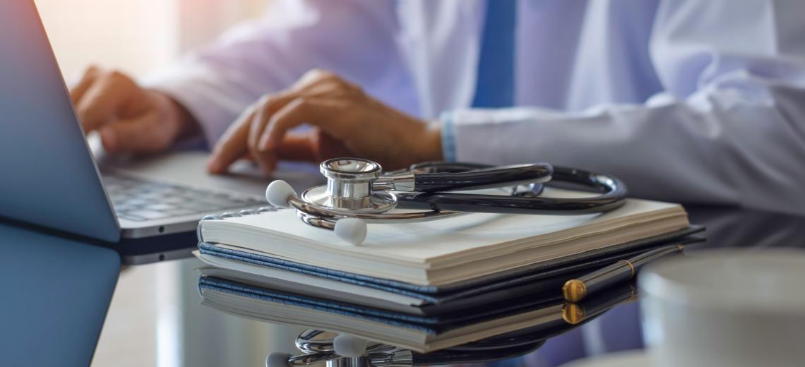 Female doctor in white lab coat typing on laptop computer with notebook and medical stethoscope on the desk at workplace. 